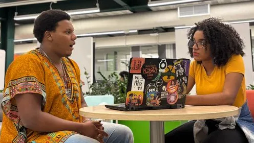 Dois jovens conversando em uma mesa com um notebook coberto de adesivos