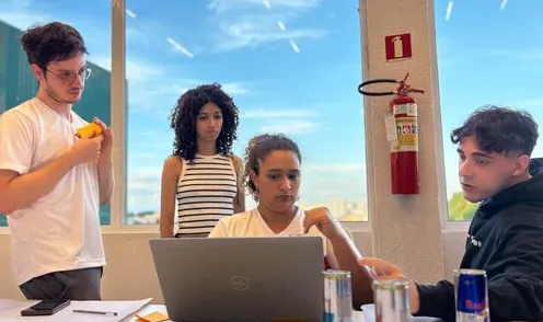 Quatro jovens reunidos em volta de um notebook em uma sala com vista para a cidade