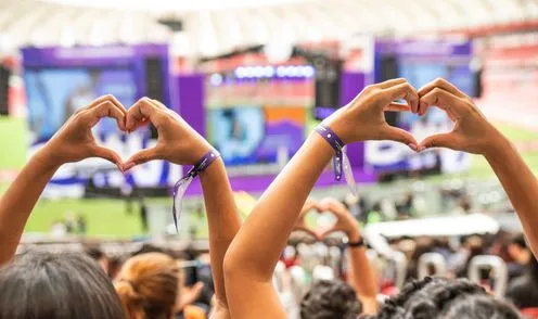 Público em um estádio fazendo o gesto de coração com as mãos