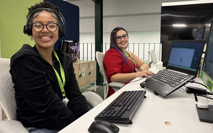 Duas jovens sorrindo em suas estações de trabalho, com computadores sobre a mesa